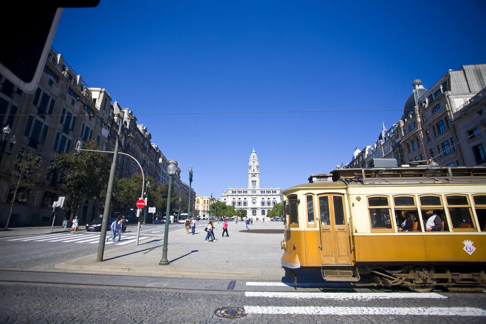 A yellow tram crosses a wide Porto street, with the clock tower of City Hall at the far end under a deep blue sky