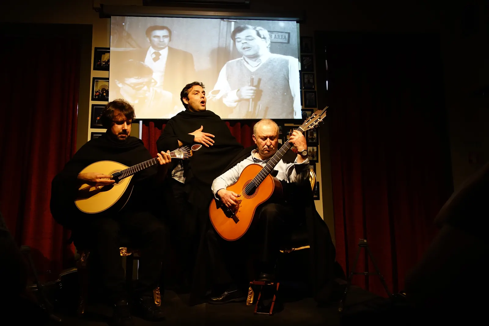 Three men in black academic capes on a stage with red curtains, two playing guitars and one singing, with a black-and-white film projected behind them