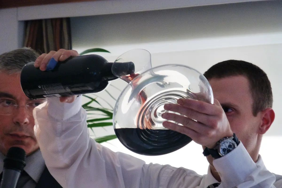 A man tips a dark bottle of port into a large glass decanter held up to the light