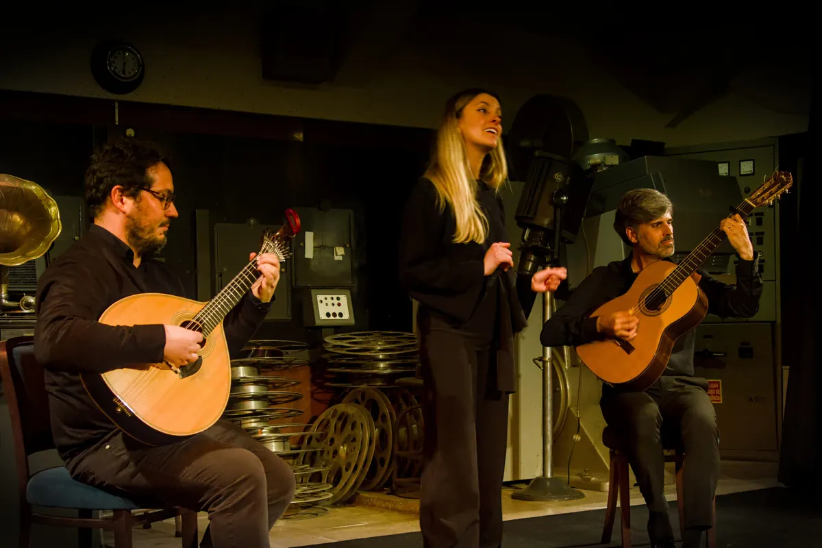 A fado singer standing between two seated guitarists, one with a Portuguese guitar and one with a classical guitar